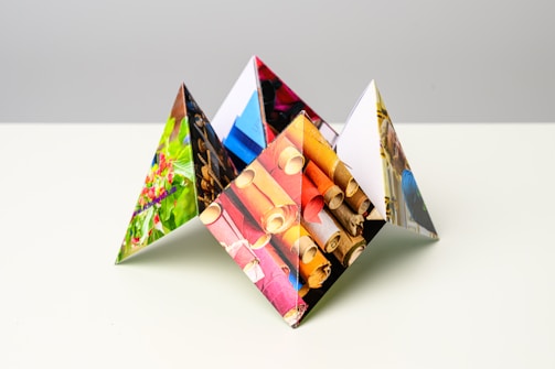 An abstract geometric sculpture composed of triangular shapes, each face featuring different vibrant photographic imagery. The images include various subjects, such as textiles, flowers, and vivid colors, creating a visually stimulating composition on a neutral light background.