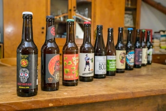 A row of different beer bottles is lined up on a wooden table, each featuring unique and colorful labels. The setting appears to be indoors with wooden cabinets in the background, adding to a rustic ambiance.
