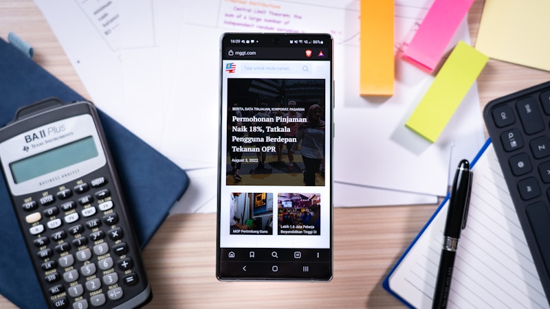 A smartphone displaying a news website rests on a wooden desk surrounded by office supplies. The desk includes a calculator, colorful sticky notes, sheets of paper, a pen, and the edge of a keyboard. The setting suggests a workspace or study area.