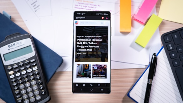 A smartphone displaying a news website rests on a wooden desk surrounded by office supplies. The desk includes a calculator, colorful sticky notes, sheets of paper, a pen, and the edge of a keyboard. The setting suggests a workspace or study area.