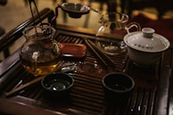 On a wooden tea tray, there are various tea implements including a glass teapot with a golden liquid, a white ceramic gaiwan with inscriptions, a stainless steel strainer, and other small tea cups and utensils. The setup suggests a traditional tea brewing session with a focus on Chinese tea culture.