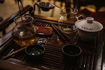 On a wooden tea tray, there are various tea implements including a glass teapot with a golden liquid, a white ceramic gaiwan with inscriptions, a stainless steel strainer, and other small tea cups and utensils. The setup suggests a traditional tea brewing session with a focus on Chinese tea culture.