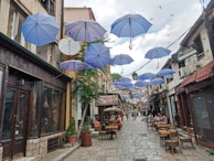 A narrow cobblestone street in a quaint urban area, with blue umbrellas suspended overhead. The street is lined with small shops, cafes, and restaurants. People are seated at tables outside the cafes, and others are walking along the street. Buildings have a mix of stone and wooden facades with various signs.