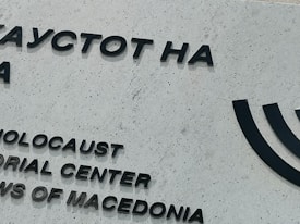 A portion of a building wall featuring black text in both Cyrillic and English. The text reads 'Holocaust Memorial Center for the Jews of Macedonia.' The wall is light gray and textured, and there is a partial image of a Menorah symbol to the right.