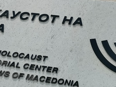A portion of a building wall featuring black text in both Cyrillic and English. The text reads 'Holocaust Memorial Center for the Jews of Macedonia.' The wall is light gray and textured, and there is a partial image of a Menorah symbol to the right.
