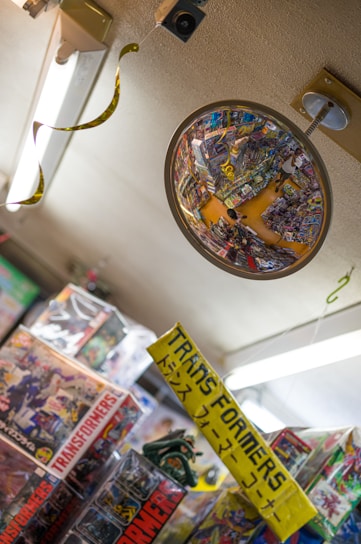 A convex security mirror reflects a cluttered toy store aisle filled with colorful merchandise, primarily focused on Transformers toys. Numerous toy boxes and a bright yellow sign with both English and Japanese text are visible. The ceiling has fluorescent lighting and some hanging decorations.