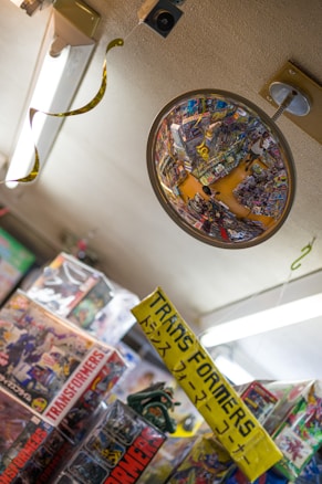 A convex security mirror reflects a cluttered toy store aisle filled with colorful merchandise, primarily focused on Transformers toys. Numerous toy boxes and a bright yellow sign with both English and Japanese text are visible. The ceiling has fluorescent lighting and some hanging decorations.