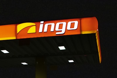 A brightly illuminated gas station canopy at night with the logo 'ingo' displayed prominently in white and a partially yellow background on an orange panel.