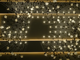 A ceiling installation featuring numerous small lights radiating from the center, resembling a floral pattern. Metallic flower-like structures are interspersed among the lights, creating a dazzling and dynamic visual effect.