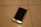 A smartphone with a cracked screen is lying on a tiled floor. The cracks are prominently visible on the screen, which is black, and the phone appears to be placed at an angle. The tile floor is light-colored with visible grout lines.
