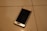 A smartphone with a cracked screen is lying on a tiled floor. The cracks are prominently visible on the screen, which is black, and the phone appears to be placed at an angle. The tile floor is light-colored with visible grout lines.
