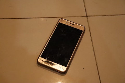 A smartphone with a cracked screen is lying on a tiled floor. The cracks are prominently visible on the screen, which is black, and the phone appears to be placed at an angle. The tile floor is light-colored with visible grout lines.