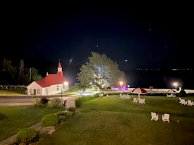 A serene nighttime scene featuring a small church with a red roof and white walls, situated near a quiet road. A large, illuminated tree stands nearby, casting a soft glow over the area. The open grassy space is dotted with white chairs and umbrellas, suggesting a peaceful outdoor setting.
