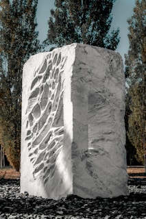 A large, textured stone sculpture with intricate carvings occupies the center, surrounded by tall, slender trees. The sculpture is made of light-colored stone, featuring a mix of smooth surfaces and deep grooves. The setting is outdoors on a layer of dark stones, with sunlight highlighting the details of the sculpture.