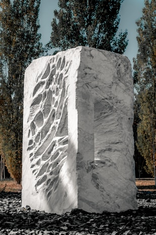 A large, textured stone sculpture with intricate carvings occupies the center, surrounded by tall, slender trees. The sculpture is made of light-colored stone, featuring a mix of smooth surfaces and deep grooves. The setting is outdoors on a layer of dark stones, with sunlight highlighting the details of the sculpture.