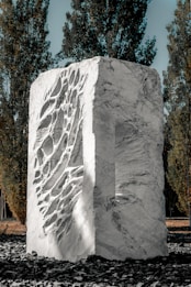 A large, textured stone sculpture with intricate carvings occupies the center, surrounded by tall, slender trees. The sculpture is made of light-colored stone, featuring a mix of smooth surfaces and deep grooves. The setting is outdoors on a layer of dark stones, with sunlight highlighting the details of the sculpture.