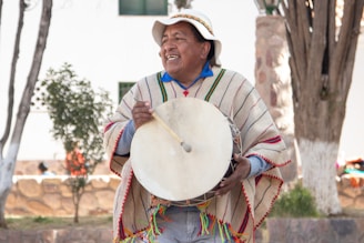 A person wearing a colorful poncho and a traditional hat is playing a drum outdoors. The setting appears to be a park or garden with trees and a stone wall in the background. The person is smiling, indicating a cheerful atmosphere.