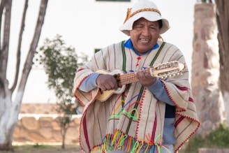A person wearing traditional colorful clothing and a hat is playing a stringed instrument outdoors. The setting includes trees and a stone structure in the background, suggesting a natural or rustic environment.