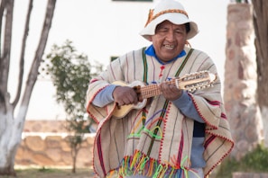 A person wearing traditional colorful clothing and a hat is playing a stringed instrument outdoors. The setting includes trees and a stone structure in the background, suggesting a natural or rustic environment.