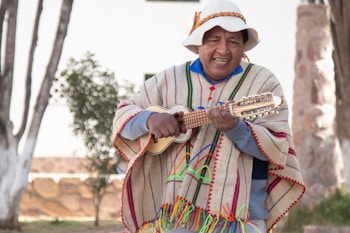 A person wearing traditional colorful clothing and a hat is playing a stringed instrument outdoors. The setting includes trees and a stone structure in the background, suggesting a natural or rustic environment.