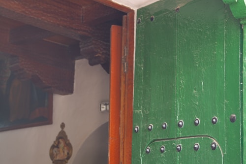 A green wooden door partially open, revealing an interior space with wooden ceiling beams. There is a wall decoration with an ornate design and a framed painting in the background. The door has metal studs and detailing.