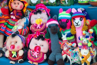 A collection of colorful handmade dolls and stuffed animals displayed together. The dolls are dressed in vibrant traditional attire, featuring bright colors and patterns. Several plush llamas and other animals knitted in bright woolen textures are also present, creating a lively and festive appearance.