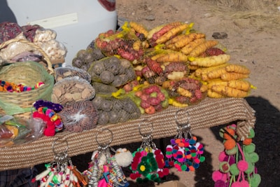 A display of various types of potatoes in nets placed on a table. There are also colorful pom-pom keychains hanging from the table edge. A basket containing more pom-poms is visible alongside several bags filled with dry goods. The setup is on an outdoor surface with patches of grass and dirt.