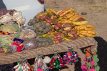 A display of various types of potatoes in nets placed on a table. There are also colorful pom-pom keychains hanging from the table edge. A basket containing more pom-poms is visible alongside several bags filled with dry goods. The setup is on an outdoor surface with patches of grass and dirt.