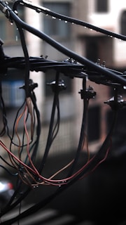 A clustered arrangement of electrical wires suspended outdoors against a blurred urban background. The wires are bundled and tangled, with some insulation stripped, revealing colorful inner wires. There are metallic clamps securing the wires to a horizontal bar. It's a cloudy day with visible droplets of rain.