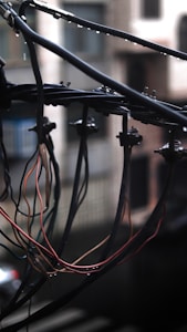 A clustered arrangement of electrical wires suspended outdoors against a blurred urban background. The wires are bundled and tangled, with some insulation stripped, revealing colorful inner wires. There are metallic clamps securing the wires to a horizontal bar. It's a cloudy day with visible droplets of rain.