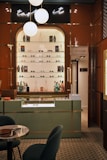 A stylish eyewear store interior featuring a wall shelf with a variety of eyeglasses and decorative items. Above the shelf, a neon sign glows softly with the words 'taste it.' The wall panels are a warm brown color, contrasting with the sleek, modern aesthetic. The foreground includes a display counter with a glass top, set against a patterned tiled floor. A cozy seating arrangement with a small table is visible on the left, and a door labeled 'Opticalab' is on the right side of the image.