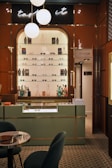A stylish eyewear store interior featuring a wall shelf with a variety of eyeglasses and decorative items. Above the shelf, a neon sign glows softly with the words 'taste it.' The wall panels are a warm brown color, contrasting with the sleek, modern aesthetic. The foreground includes a display counter with a glass top, set against a patterned tiled floor. A cozy seating arrangement with a small table is visible on the left, and a door labeled 'Opticalab' is on the right side of the image.