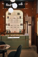 A stylish eyewear store interior featuring a wall shelf with a variety of eyeglasses and decorative items. Above the shelf, a neon sign glows softly with the words 'taste it.' The wall panels are a warm brown color, contrasting with the sleek, modern aesthetic. The foreground includes a display counter with a glass top, set against a patterned tiled floor. A cozy seating arrangement with a small table is visible on the left, and a door labeled 'Opticalab' is on the right side of the image.