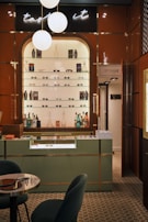 A stylish eyewear store interior featuring a wall shelf with a variety of eyeglasses and decorative items. Above the shelf, a neon sign glows softly with the words 'taste it.' The wall panels are a warm brown color, contrasting with the sleek, modern aesthetic. The foreground includes a display counter with a glass top, set against a patterned tiled floor. A cozy seating arrangement with a small table is visible on the left, and a door labeled 'Opticalab' is on the right side of the image.