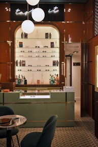 A stylish eyewear store interior featuring a wall shelf with a variety of eyeglasses and decorative items. Above the shelf, a neon sign glows softly with the words 'taste it.' The wall panels are a warm brown color, contrasting with the sleek, modern aesthetic. The foreground includes a display counter with a glass top, set against a patterned tiled floor. A cozy seating arrangement with a small table is visible on the left, and a door labeled 'Opticalab' is on the right side of the image.