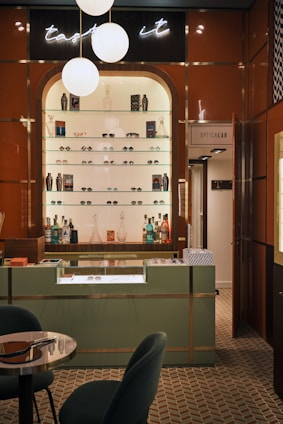 A stylish eyewear store interior featuring a wall shelf with a variety of eyeglasses and decorative items. Above the shelf, a neon sign glows softly with the words 'taste it.' The wall panels are a warm brown color, contrasting with the sleek, modern aesthetic. The foreground includes a display counter with a glass top, set against a patterned tiled floor. A cozy seating arrangement with a small table is visible on the left, and a door labeled 'Opticalab' is on the right side of the image.