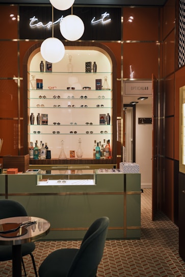 A stylish eyewear store interior featuring a wall shelf with a variety of eyeglasses and decorative items. Above the shelf, a neon sign glows softly with the words 'taste it.' The wall panels are a warm brown color, contrasting with the sleek, modern aesthetic. The foreground includes a display counter with a glass top, set against a patterned tiled floor. A cozy seating arrangement with a small table is visible on the left, and a door labeled 'Opticalab' is on the right side of the image.