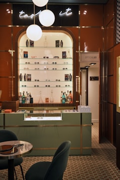 A stylish eyewear store interior featuring a wall shelf with a variety of eyeglasses and decorative items. Above the shelf, a neon sign glows softly with the words 'taste it.' The wall panels are a warm brown color, contrasting with the sleek, modern aesthetic. The foreground includes a display counter with a glass top, set against a patterned tiled floor. A cozy seating arrangement with a small table is visible on the left, and a door labeled 'Opticalab' is on the right side of the image.