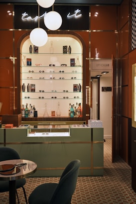 A stylish eyewear store interior featuring a wall shelf with a variety of eyeglasses and decorative items. Above the shelf, a neon sign glows softly with the words 'taste it.' The wall panels are a warm brown color, contrasting with the sleek, modern aesthetic. The foreground includes a display counter with a glass top, set against a patterned tiled floor. A cozy seating arrangement with a small table is visible on the left, and a door labeled 'Opticalab' is on the right side of the image.
