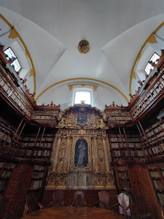 An ornate library setting with high ceilings and a large, gilded altar-like structure at the center. The walls are lined with tall bookshelves filled with books. The ceiling is arched, adorned with decorative molding, and features large windows that let in natural light. A few empty chairs are positioned in front of the central structure, adding an air of quiet reverence.