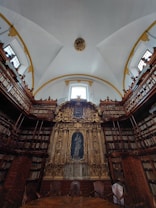 An ornate library setting with high ceilings and a large, gilded altar-like structure at the center. The walls are lined with tall bookshelves filled with books. The ceiling is arched, adorned with decorative molding, and features large windows that let in natural light. A few empty chairs are positioned in front of the central structure, adding an air of quiet reverence.