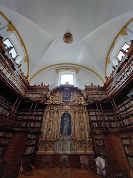 An ornate library setting with high ceilings and a large, gilded altar-like structure at the center. The walls are lined with tall bookshelves filled with books. The ceiling is arched, adorned with decorative molding, and features large windows that let in natural light. A few empty chairs are positioned in front of the central structure, adding an air of quiet reverence.