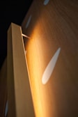 A close-up view of a wooden panel with a warm light illuminating from behind its edge. The surface of the wood features a smooth texture with glossy white teardrop-shaped patterns, creating an abstract and modern aesthetic.