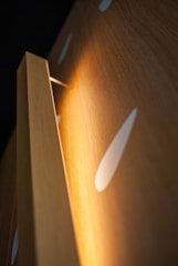 A close-up view of a wooden panel with a warm light illuminating from behind its edge. The surface of the wood features a smooth texture with glossy white teardrop-shaped patterns, creating an abstract and modern aesthetic.