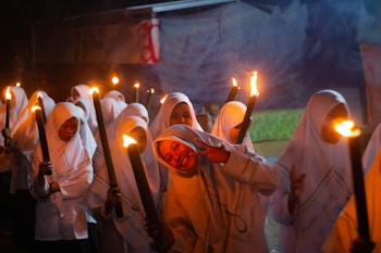 A group of people dressed in white garments are holding torches in what appears to be a night-time gathering. One person in the foreground is smiling and making a peace sign. The scene suggests a sense of camaraderie and unity.