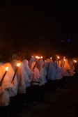 A group of people dressed in white cloaks holding torches, walking in a procession during nighttime. The bright flames from the torches create a stark contrast with the surrounding darkness, illuminating the faces partially covered by the cloaks.