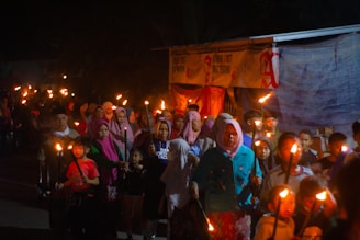 A group of people, including both adults and children, are participating in a nighttime torch march. They are holding lit torches, and many of them are wearing colorful clothing, including headscarves. The scene is dimly lit, creating a warm glow around the individuals.