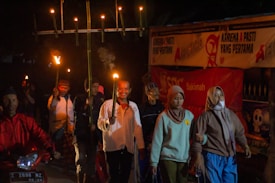 A group of people are participating in a nighttime procession, holding torches made from bamboo. They are dressed in various traditional and casual attire. Banners with writing and symbols are visible in the background.