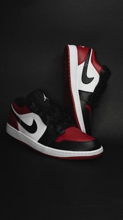 A pair of stylish athletic shoes in black, white, and red colors with distinctive branding. The shoes are positioned against a dark background, showcasing their sleek design and detailed craftsmanship.