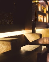 A cozy seating area with plush, cushioned benches and a polished wooden table. The ambient lighting casts a warm glow, highlighting a textured wall in the background. Shelves with decorative items are visible on the right side.